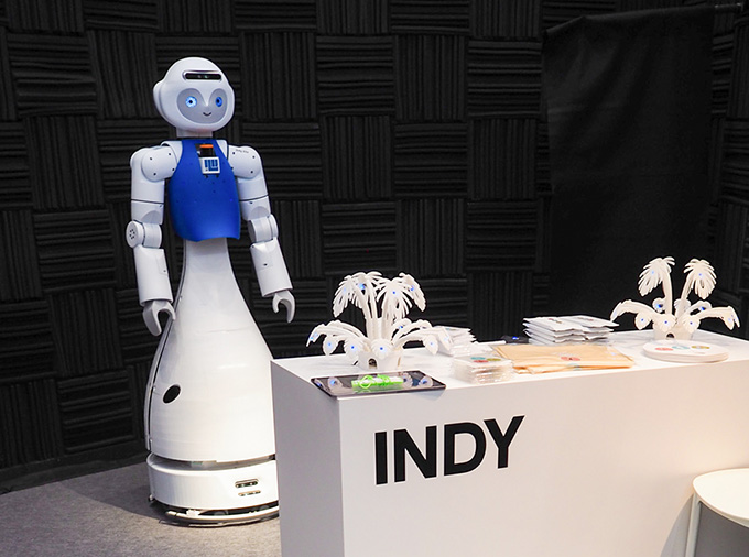 来場者から話かけられるのを待つIndy。机に並んでいるのはコースター、エコバック、団扇などのノベルティグッズの様子