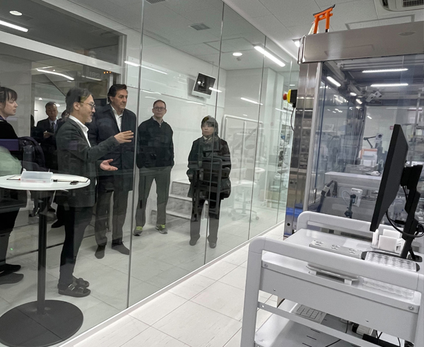 高橋 プロジェクトディレクターから実験自動化技術の説明を受けられるギル 次官の写真