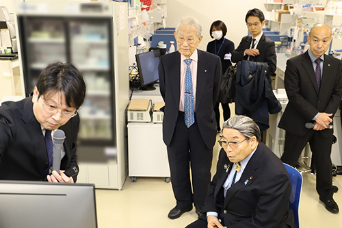 井上チームリーダー(左)から説明を受ける北村大臣(右)(後列中央は松本理事長)の写真